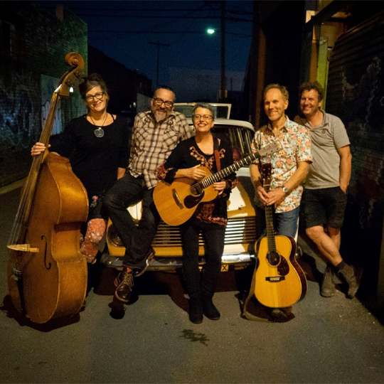A group of five people stand against a car in a back laneway at night under a lamppost holding guitars and a double bass